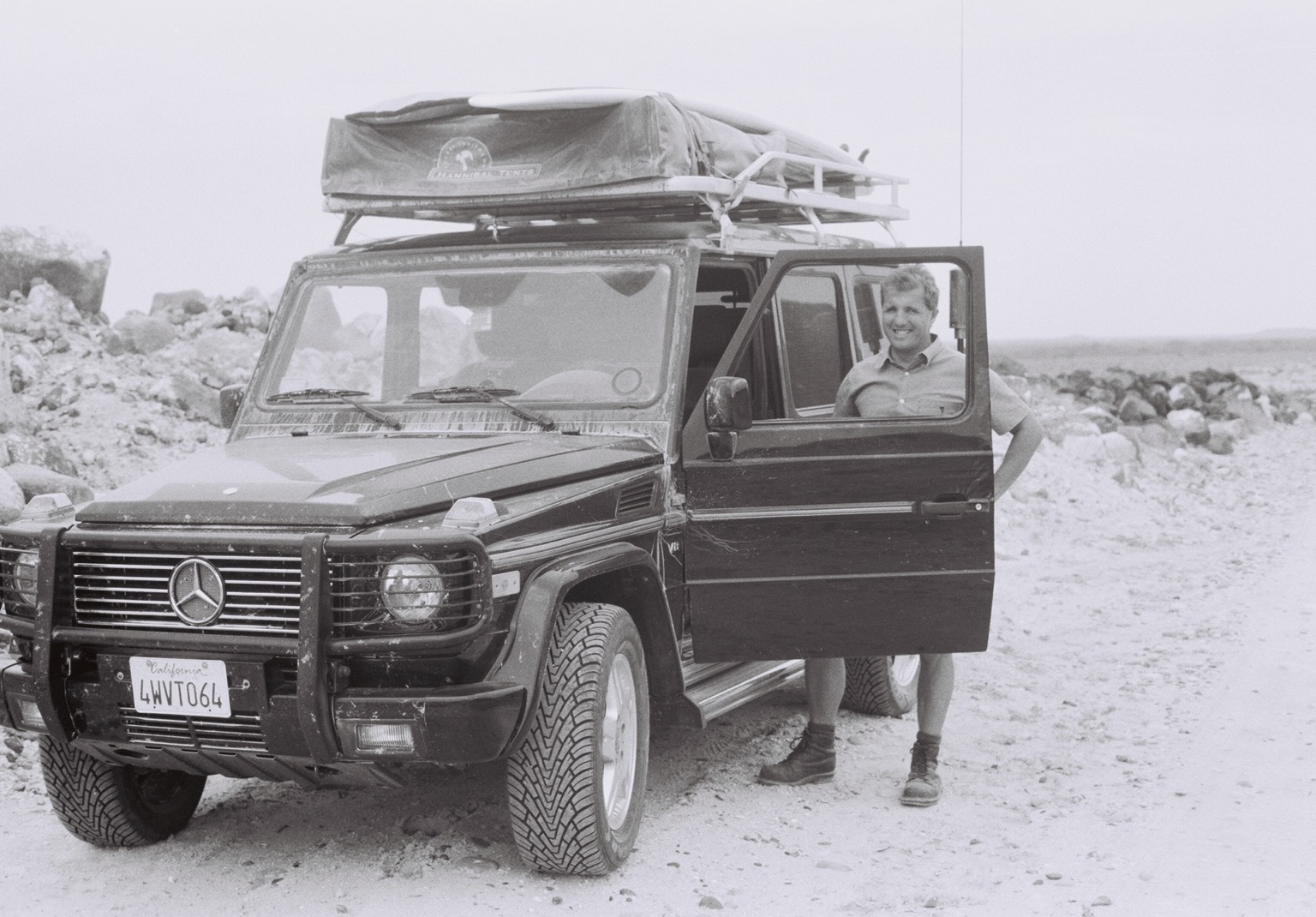 A person stands beside  black Mercedes-Benz G-Class SUV with the driver door open on a rocky, sandy terrain. The vehicle has a rooftop tent and surfboards secured on top. The license plate reads California 4WVT064 and the rooftop tent cover displays the logo Hannibal Safari Equipment. The environment appears remote and rugged, with rocks and a barren landscape in the background. The scene conveys a sense of adventure and exploration.